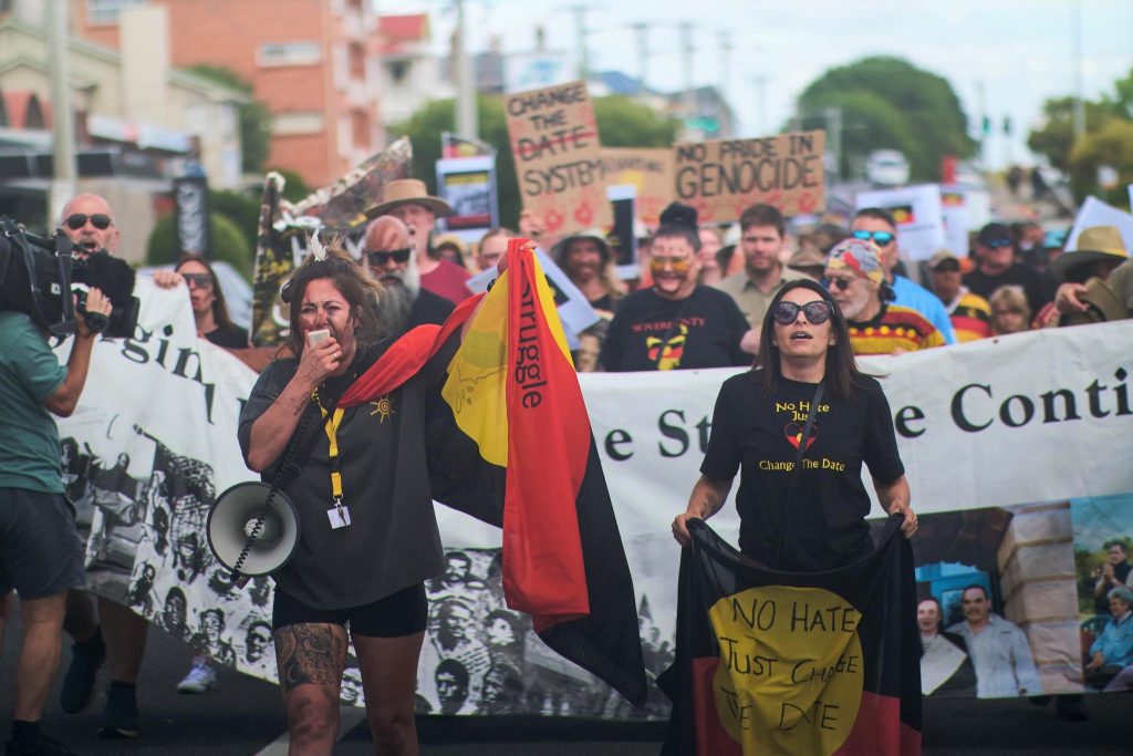 A photo of hundreds of Aborigines and supporters walking down the street with two women in front, one with a flag around her shoulders and chanting through a megaphone and the other holding a flag that says No Hate, Just Change the Date