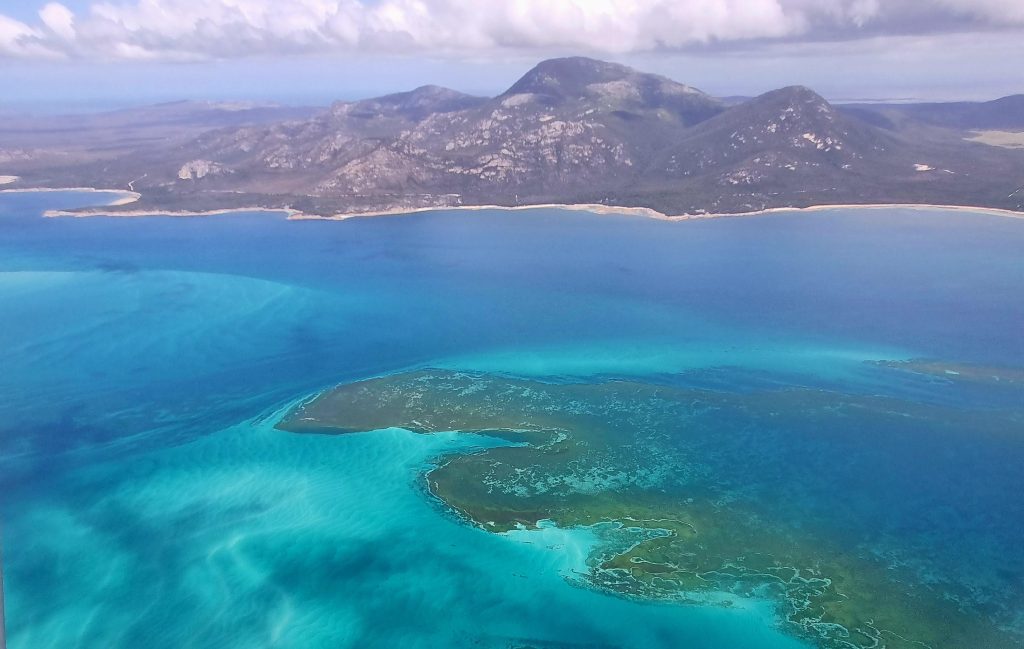 An aerial photo taken from a small plane looking over tayaritja/Bass Strait Islands. In the foreground is the turquoise waters and reefs of sea country with an island in the background rising up into the clouds.