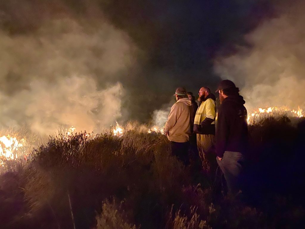 A night photo of four rangers standing side oh as they look at a cultural fire burning. Their faces are lit by the light of the fire. Flames and smoke rise up from the grasslands in front of the rangers. 