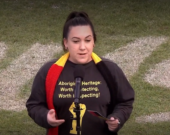 Photo of Daisy Allen, an Aboriginal woman, standing in front of a microphone on a football field. Daisy is wearing a black top that says Aboriginal Heritage: worth protecting, worth respecting, in yellow writing, with a yellow fist raised underneath. She is also wearing a black, yellow and red knitted scarf around her neck. Daisy has brown hair in a ponytail.